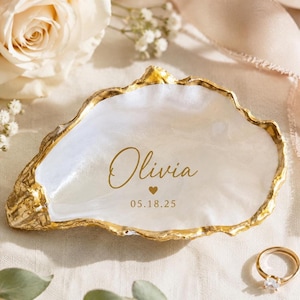 May include: A white oyster shell trinket dish with a gold-painted rim. The center features the gold text Olivia, a small heart icon, and the date 05.18.25. The interior surface has a pearlescent finish, and the exterior edge has a textured gold finish.