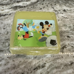 May include: A yellow plastic lunchbox with a Mickey Mouse, Donald Duck, and Goofy design. The characters are playing soccer on a green field.