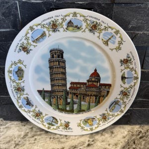 May include: A white ceramic plate with a colourful design featuring the Leaning Tower of Pisa and other Italian landmarks. The plate has a blue and yellow border with the words "Souvenir Italy" repeated around the edge. The text "Pisa - Torre Pendente e Abside del Duomo" is printed at the bottom of the plate.