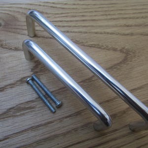 May include: Two polished chrome cabinet pulls with mounting screws on a wooden surface. The pulls are a rounded, tubular design, with a curved handle and a rectangular base. The screws are silver.