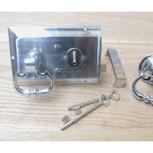 Könnte beinhalten: Ein silberfarbenes Türschloss-Set mit einem rechteckigen Schloss mit Griff, einem runden Türklopfer, einem Schließblech und zwei Schlüsseln. Das Set liegt auf einer hellen Holzoberfläche. Das Schloss ist etwa 15 cm lang.