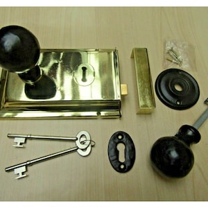 Könnte beinhalten: Ein Vintage-Türschloss-Set aus Messing mit schwarzem Knauf und passender Hardware. Das Set enthält ein rechteckiges Schloss, ein Schließblech, zwei Schlüssel und eine runde Rückplatte. Die Hardware ist auf einer hellen Holzoberfläche angeordnet.