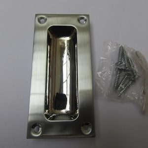 Könnte beinhalten: Ein rechteckiger, gebürsteter Nickel-Einlassgriff mit abgerundetem Innenraum. Vier Schraubenlöcher sind sichtbar. Ein kleiner Plastikbeutel enthält mehrere silberne Schrauben und ein kleines Werkzeug.