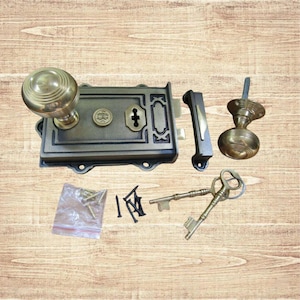 May include: An antique bronze door lock set, featuring a round doorknob, a rectangular lock plate, a latch, and two skeleton keys. The set includes screws and other hardware. The hardware is displayed on a wooden surface.
