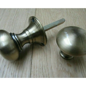 Könnte beinhalten: Zwei antike Türknöpfe aus Messing. Ein Knopf ist an einer Spindel und einer Montageplatte befestigt. Der andere Knopf ist separat. Die Knöpfe haben eine ovale Form und eine warme, metallische Oberfläche. Die Hardware liegt auf einer hellen Holzoberfläche.