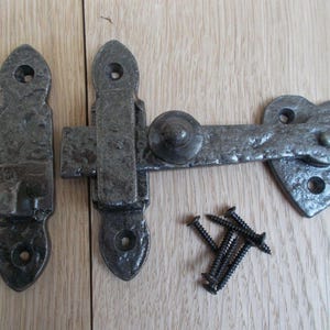 Könnte beinhalten: Ein rustikales, graues Metall-Torverschluss-Set mit herzförmigem Ende. Das Set enthält den Riegel, eine Schließplatte und einen Satz schwarzer Schrauben. Das Metall hat eine gehämmerten Textur, die ihm einen Vintage-Look verleiht.
