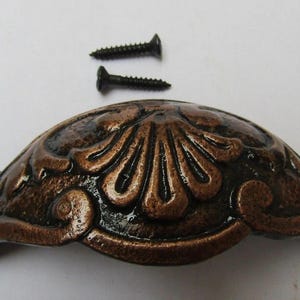 Könnte beinhalten: Verzierte, bronzefarbene Schrankgriff mit dekorativem Blumendesign und zwei schwarzen Schrauben. Der Griff hat eine gebogene Form mit Befestigungslöchern an jedem Ende, geeignet für Möbelrestaurierung oder Neubauten.