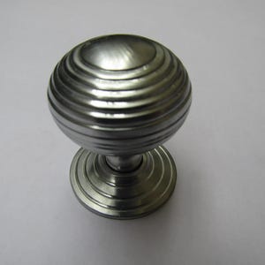 May include: A silver-toned cabinet knob with a round, ribbed design. The knob sits on a circular base with concentric rings. This hardware piece is suitable for drawers, cabinets, or furniture.