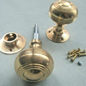 Könnte beinhalten: Ein Satz Messing-Türknöpfe mit rundem, gerilltem Design. Das Set enthält zwei Knöpfe, eine Montageplatte, eine Verbindungsstange und mehrere Schrauben. Die Hardware hat eine warme Goldfarbe.