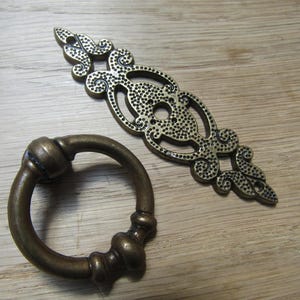 Puede incluir: Dos herrajes de gabinete antiguos de color bronce. Uno es un tirador de anillo con un diseño de nudo decorativo, y el otro es una placa trasera ornamentada con un patrón punteado. El herraje está sobre una superficie de madera clara.