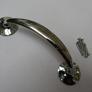 Puede incluir: Un tirador de armario cromado brillante con un diseño curvo. El tirador tiene dos placas de montaje circulares con orificios para tornillos. Un pequeño montón de tornillos plateados está al lado del tirador.