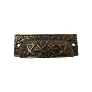 May include: A bronze-colored drawer pull with an ornate, raised design. The rectangular pull features a central recessed area with geometric and floral patterns, and two holes for mounting. The edges are detailed with a floral motif.