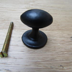 Könnte beinhalten: Schwarzer, ovaler Schrankknauf mit passendem Sockel und goldfarbener Schraube. Der Knauf hat eine glatte, matte Oberfläche. Die Schraube ist ca. 5 cm lang. Möbelbeschläge.