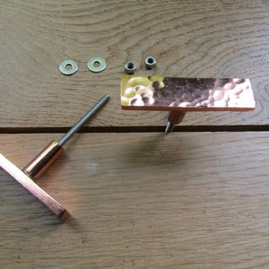 Könnte beinhalten: Zwei kupferfarbene Schrankgriffe mit gehammerter Textur, zusammen mit Unterlegscheiben und Muttern, sind auf einer Holzoberfläche angeordnet. Ein Griff ist rechteckig, der andere quadratisch. Möbelbeschläge.