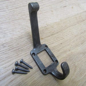 Könnte beinhalten: Ein dunkelgrauer Metallhaken mit quadratischer Öffnung und gebogenem Haken. Vier Schrauben liegen neben dem Haken. Der Haken ist zum Aufhängen von Gegenständen konzipiert und hat einen rustikalen, industriellen Stil.