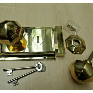 Könnte beinhalten: Ein Vintage-Türschloss-Set aus Messing mit zwei Schlüsseln, einem facettierten Türknauf und einem passenden Schließblech. Das Set enthält außerdem eine separate Rosette und eine kleine Packung Schrauben. Die Hardware hat eine polierte, reflektierende Oberfläche.