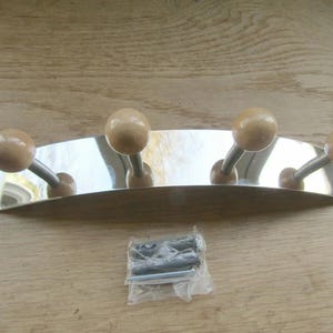 Op de afbeelding: Een verchroomde wandkapstok met vier houten bolvormige haken. De kapstok heeft een gebogen ontwerp en bevat montagemateriaal. De haken zijn gelijkmatig verdeeld over de chromen stang. De houten bollen zijn lichtbruin van kleur.
