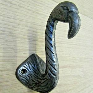 Könnte beinhalten: Ein Haken aus Gusseisen in Form eines Flamingos mit strukturierter, verwitterter Oberfläche. Der Haken hat einen detaillierten Flamingo-Kopf und -Hals mit einem dekorativen, geriffelten Design am Hals. Die Basis ist rautenförmig.