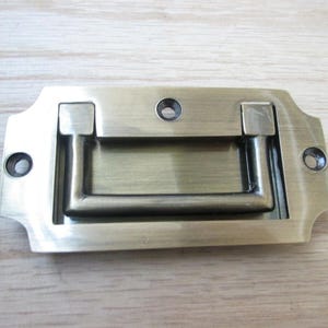 Puede incluir: Tirador de cajón de latón antiguo con un asa rectangular y placa trasera decorativa. El tirador presenta un asa empotrada y dos orificios para tornillos a cada lado para el montaje. Una pieza de ferretería clásica.
