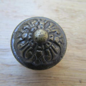 Könnte beinhalten: Nahaufnahme eines runden, antiken Messing-Schrankknaufs. Der Knauf weist ein detailliertes Blumendesign mit einem zentralen, erhabenen Kreis auf. Die Oberfläche hat ein leicht gealtertes Aussehen, was auf Vintage- oder rustikale Dekoration hindeutet.