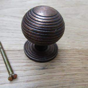 Könnte beinhalten: Ein bronzefarbener, kugelförmiger Schrankknauf mit horizontalen Rillen, montiert auf einem kreisförmigen Sockel. Eine passende Schraube befindet sich links. Diese Hardware ist für Schubladen und Schränke geeignet.