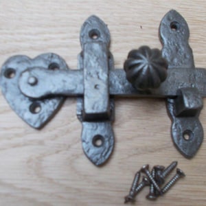 Könnte beinhalten: Ein rustikaler Torverschluss aus Gusseisen mit herzförmiger Grundplatte. Der Verschluss verfügt über einen dekorativen Knauf und wird mit mehreren Schrauben geliefert. Das Metall hat ein strukturiertes, gealtertes Aussehen.