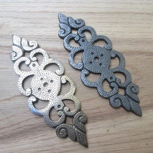 Könnte beinhalten: Zwei verzierte Metallbeschläge mit silbernem Finish. Jedes Teil hat ein symmetrisches, wirbelndes Design mit einem zentralen kreisförmigen Element und mehreren Löchern. Die Beschläge werden auf einer hellen Holzoberfläche präsentiert.