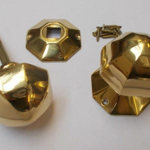 Könnte beinhalten: Ein Satz polierter Messing-Türbeschläge, darunter zwei facettierte Türknöpfe, eine quadratische Rückplatte und Schrauben. Die Beschläge haben ein geometrisches Design und eine glänzende, reflektierende Oberfläche.