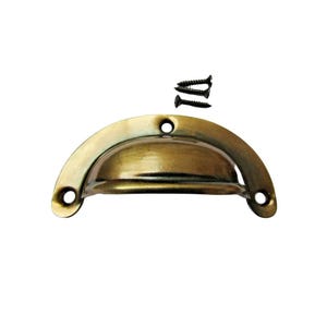 Puede incluir: Un tirador de latón de estilo vintage con tres tornillos negros. El tirador tiene un diseño semicircular con dos orificios de montaje. El acabado de latón tiene un aspecto envejecido, lo que sugiere una estética clásica o antigua.