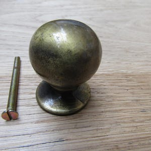 Könnte beinhalten: Ein Möbelknopf aus Messing im Vintage-Stil mit kugelförmigem Design. Der Knopf sitzt auf einem kleinen, kreisförmigen Sockel und wird von einer passenden Schraube begleitet. Die Hardware hat eine angelaufene, antike Oberfläche. Der Knopf ist wahrscheinlich für Möbel.