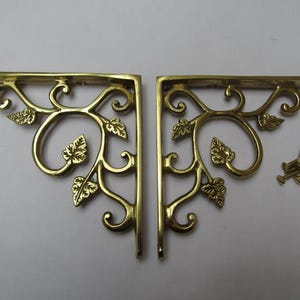 Puede incluir: Dos escuadras de estantería de latón con un diseño decorativo de hojas y vides. Las escuadras tienen forma de L con una parte superior plana y un soporte vertical. Se incluyen pequeños tornillos de latón. Las escuadras son de color dorado cálido.