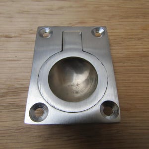 Puede incluir: Un tirador empotrado de metal cepillado con un asa circular y una base rectangular. El asa está diseñada para retraerse en la base. La base tiene cuatro orificios para tornillos para el montaje. El herraje es de color plateado.