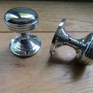 Könnte beinhalten: Zwei glänzende, verchromte Türknöpfe. Ein Knopf steht aufrecht, der andere liegt auf der Seite. Die Knöpfe haben ein rundes, geriffeltes Design und liegen auf einer hellbraunen Holzoberfläche.