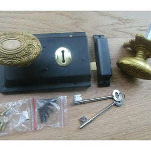 Könnte beinhalten: Antikes Türschloss-Set mit schwarzem Metallgehäuse, Messing-Schlüsselloch und verzierten Messinggriffen. Enthält einen Riegel, Schließblech, Schlüssel und Befestigungsmaterial. Die Hardware wird auf einer Holzoberfläche präsentiert.