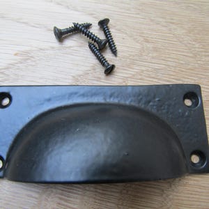 May include: Black metal cup pull with four screw holes and four black screws. The cup pull has a rounded, semi-circular design and a textured surface. Hardware for cabinets and drawers.