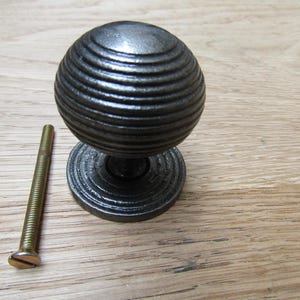 Könnte beinhalten: Ein dunkelgrauer, geriffelter Metalltürknauf mit passendem Sockel und einer langen, goldfarbenen Schraube. Der Knauf hat eine kugelförmige Form mit horizontalen Rillen. Die Hardware befindet sich auf einer hellbraunen Holzoberfläche.