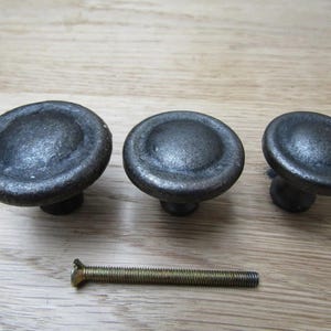 Könnte beinhalten: Drei dunkelgraue Schrankknöpfe aus Gusseisen mit passender Schraube. Die Knöpfe haben eine abgerundete, flache Oberseite und einen kurzen, zylindrischen Sockel. Die Hardware wird auf einer hellen Holzoberfläche präsentiert.