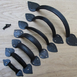 Może przedstawiać: Zestaw czarnych, kutych żelaznych uchwytów w różnych rozmiarach, ułożonych na jasnej drewnianej powierzchni. Każdy uchwyt ma zakrzywiony uchwyt i ozdobne, liściaste płytki montażowe z wstępnie wywierconymi otworami. Widoczne są również śruby.