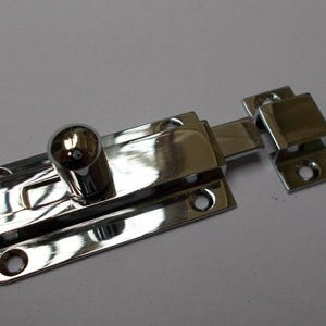 Könnte beinhalten: Ein glänzender, verchromter Türriegel mit rundem Knauf und rechteckiger Basis. Der Riegel verfügt über einen Schiebemechanismus und ist für einen sicheren Verschluss konzipiert. Inklusive Befestigungsmaterial.