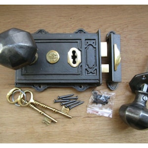 Könnte beinhalten: Antikes Türbeschlagsset, bestehend aus einem schwarzen Metallschloss mit Messingakzenten, zwei passenden Türknöpfen, Schlüsseln und Schrauben. Das Set ist für eine Vintage- oder rustikale Ästhetik konzipiert. Das Schloss misst ca. 15 cm in der Länge.