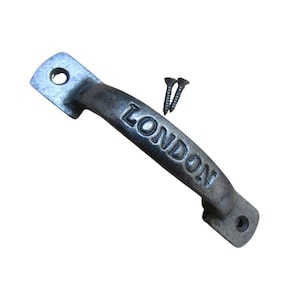 Puede incluir: Un tirador de metal plateado con la palabra "LONDON" grabada en el centro curvado. Se incluyen dos tornillos. El tirador tiene forma rectangular con bordes redondeados y agujeros para el montaje.