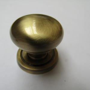 Könnte beinhalten: Nahaufnahme eines messingfarbenen Schrankknaufs. Der Knauf hat eine abgerundete, ovale Form und eine kreisförmige Basis. Das Metall hat eine gebürstete Oberfläche, die ihm ein leicht strukturiertes Aussehen verleiht. Der Knauf ist wahrscheinlich für Möbel oder Schränke.