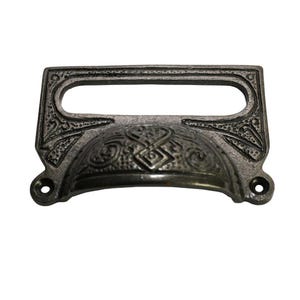 以下が含まれることがあります： 暗い灰色の仕上げが施された、装飾的なアンティークスタイルの引き出しプル。楕円形のハンドルと、装飾的なエンボス加工の花柄と幾何学模様が特徴です。プルの両側に2つのネジ穴が見えます。