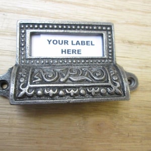 May include: A decorative metal label holder with the text "YOUR LABEL HERE" on a white insert. The holder has an ornate, vintage design with scrollwork and a dark, aged finish. It features two holes for mounting.