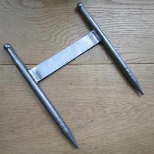 Puede incluir: Una herramienta metálica plateada con un diseño en forma de T. Cuenta con dos mangos cilíndricos con extremos puntiagudos y una pieza de conexión plana y rectangular. Los mangos tienen ranuras cerca de los extremos puntiagudos.