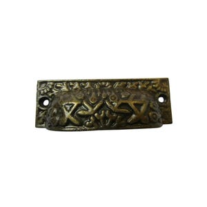 May include: Antique brass-toned drawer pull with an ornate floral and geometric design. The pull has a curved, cup-like shape with two mounting holes. This decorative hardware adds a vintage touch to furniture.