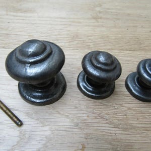 Könnte beinhalten: Drei antike, dunkelgraue Metallschrankknöpfe mit einer Schraube. Die Knöpfe haben ein abgerundetes, abgestuftes Design. Die Knöpfe sind in verschiedenen Größen erhältlich, was auf ein Set für verschiedene Schranktüren oder Schubladen hindeutet.