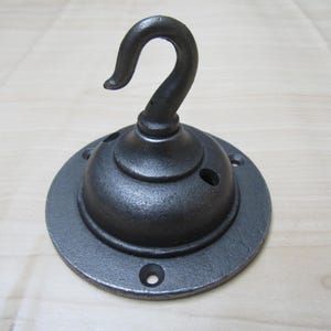 May include: A dark gray, cast iron ceiling hook with a curved hook and a circular base. The base has several holes for mounting. The hook is designed for hanging items and has a rustic, industrial aesthetic.