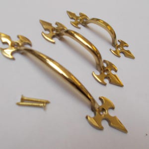 Könnte beinhalten: Ein Satz von drei goldfarbenen Schrankgriffen mit dekorativen pfeilförmigen Enden. Zwei kleine Schrauben sind ebenfalls enthalten. Die Griffe sind gebogen und haben eine glänzende Oberfläche, geeignet für Möbelrestaurierung oder Neubauten.