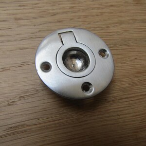 Puede incluir: Un tirador empotrado circular de metal cepillado con un centro empotrado y una lengüeta rectangular con bisagras. El tirador tiene tres pequeños orificios para tornillos y está colocado sobre una superficie de madera.